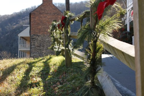 A festive garland with red ribbons decorates a wooden fence, with a hillside and building in the background.