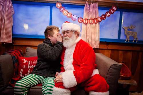 A child whispers to Santa Claus on a cozy couch, with festive decorations and a "Merry Christmas" banner in the background.