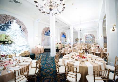 Elegant banquet hall set for an event, featuring round tables, floral arrangements, and a decorated white Christmas tree.