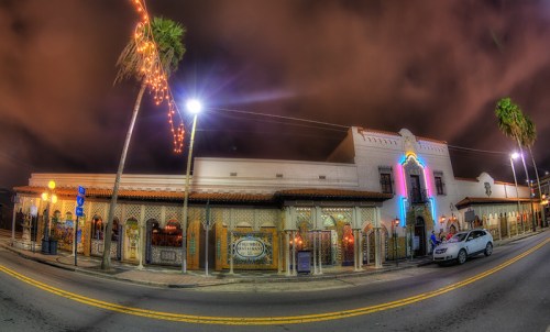 A vibrant building with neon lights and palm trees, illuminated at night along a city street.