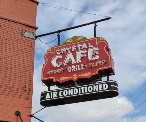 Sign for Crystal Cafe with the slogan "Never Grill, Sleep" and "Air Conditioned" below, against a cloudy sky.