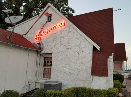 A white stone building with a red roof and neon sign reading "Herman's" at dusk.