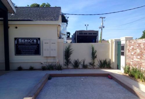 A bocce ball court with a sign, surrounded by plants and a building in the background under a clear blue sky.