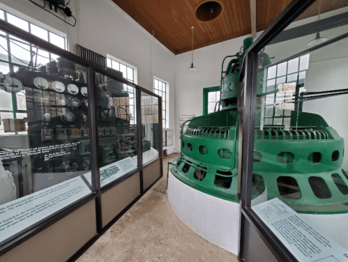 Interior of a historic power station, featuring green machinery and glass displays of electrical equipment.