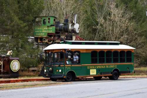 A green and yellow trolley bus parked near a vintage steam train in a scenic outdoor setting.