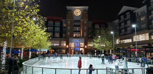 A nighttime ice skating rink surrounded by trees and buildings, with people skating and festive lights.