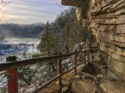 A scenic view of a river waterfall from a rocky trail, surrounded by trees and cliffs under a soft morning light.