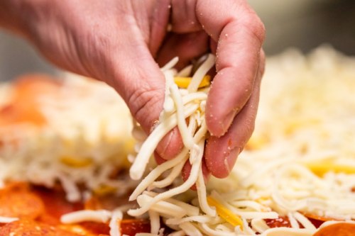 A hand sprinkles shredded cheese over a pizza topped with sauce and pepperoni.