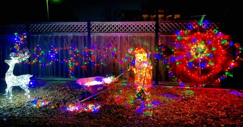 A festive scene with a lit-up Santa and reindeer, surrounded by colorful Christmas lights against a wooden fence.