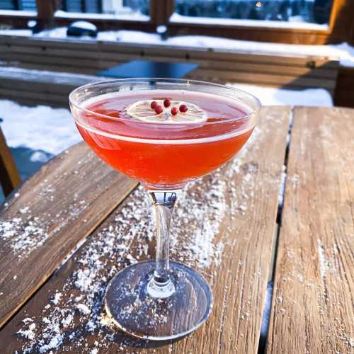 A vibrant red cocktail garnished with a lemon slice and pink peppercorns, served in a stylish glass on a snowy table.