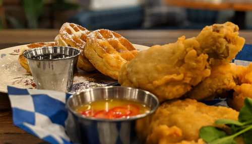A plate featuring fried chicken, waffles, and syrup, garnished with powdered sugar and a side of dipping sauce.