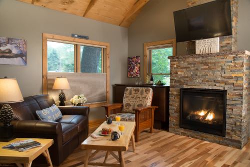 Cozy living room with a stone fireplace, wooden furniture, and large windows, featuring warm lighting and a welcoming atmosphere.