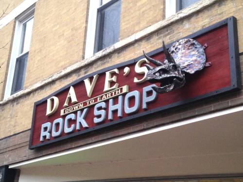 Sign for "Dave's Rock Shop" featuring a dinosaur skull design, set against a brick building backdrop.