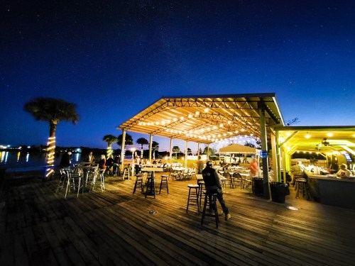A lively outdoor restaurant at night, illuminated by string lights, with tables and a view of the water.