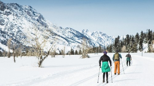 Three people cross-country skiing on a snowy trail with mountains in the background.