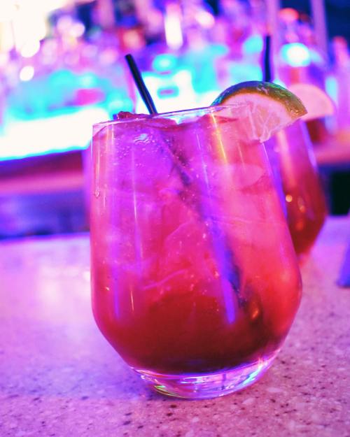 A colorful cocktail in a glass with ice, garnished with a lime wedge, set against a vibrant bar background.