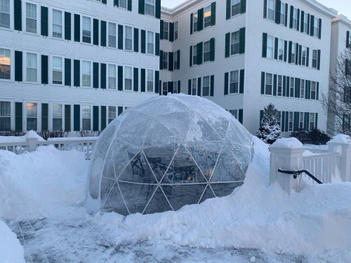 A clear geodesic dome surrounded by snow, set against a white building with green shutters.