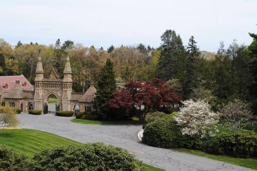 A scenic view of a stone archway surrounded by lush trees and blooming flowers on a cloudy day.