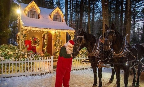 Santa in a red suit stands near a snowy house, with two horses in harness, surrounded by festive decorations.