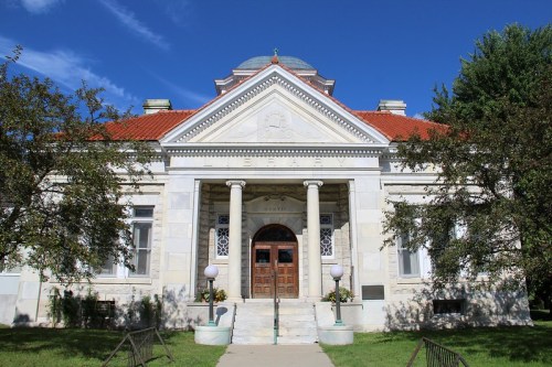 Historic building with a stone facade, columns, and a red-tiled roof, surrounded by greenery under a blue sky.