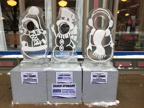 Three ice sculptures of a snowman, a penguin, and a woman in winter attire, displayed outside a building with sponsor signs.