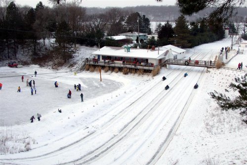 A snowy landscape with people sledding down a hill and others playing in the snow near a building.