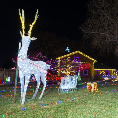 A festive yard display featuring illuminated reindeer, a decorated house, and colorful holiday lights.
