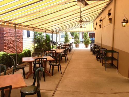Outdoor dining area with wooden tables and chairs, shaded by a striped awning, lined with potted plants.