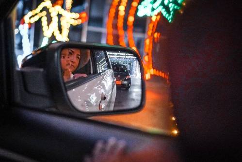 A reflection in a car mirror shows two people looking out, with colorful holiday lights in the background.