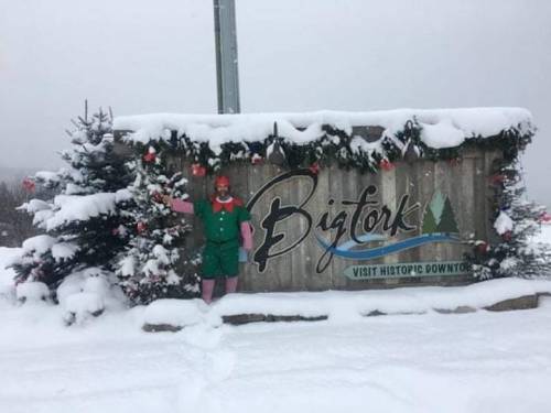 A person in a festive outfit stands next to a snow-covered sign for Bigfork, decorated with holiday ornaments.