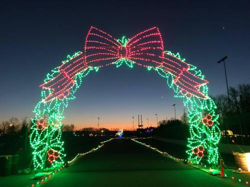A colorful holiday light arch with a bow, set against a twilight sky, leading down a festive pathway.