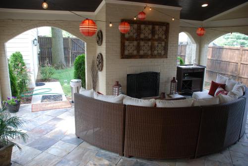 Cozy outdoor patio with a curved sofa, lanterns, and a fireplace, surrounded by greenery and a wooden fence.