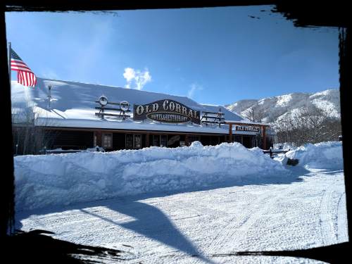 A rustic restaurant named "Old Corral" surrounded by snow, with mountains and a clear blue sky in the background.