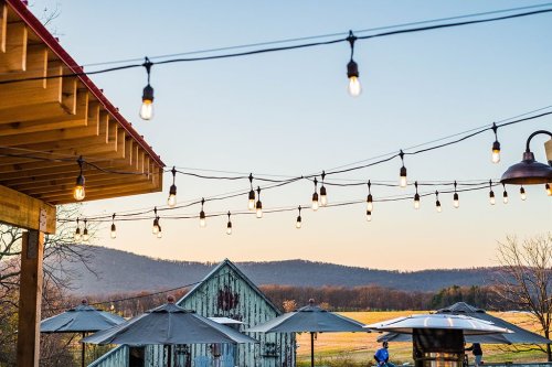 String lights hang above a patio with umbrellas, overlooking a scenic landscape and mountains in the background.