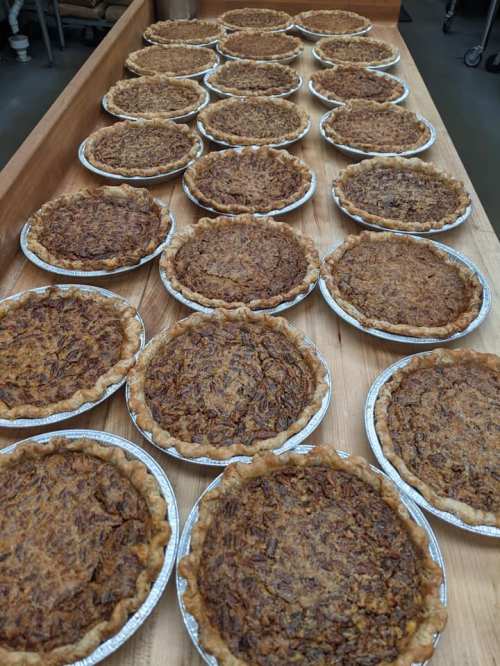 A long wooden table filled with multiple pecan pies in silver foil pans, all freshly baked and golden brown.
