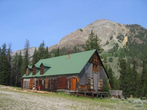 A rustic wooden cabin with a green roof, surrounded by trees and mountains in the background.