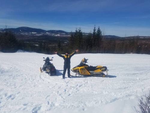 A person in winter gear stands with arms raised beside two snowmobiles on a snowy landscape with mountains in the background.