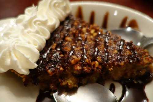 A slice of dessert topped with whipped cream and drizzled with chocolate sauce, served on a plate with a spoon.