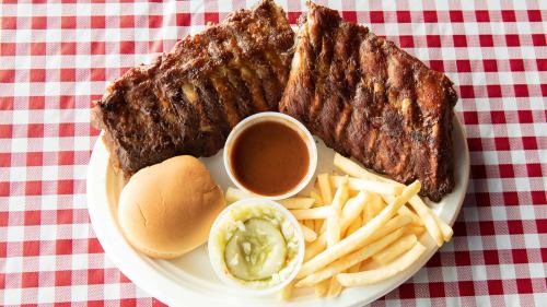 A plate of barbecue ribs, fries, a bun, and a small bowl of sauce on a red and white checkered tablecloth.