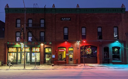 A brick building with colorful lights, snow on the ground, and storefronts visible at dusk.