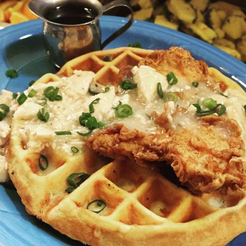 A golden waffle topped with fried chicken, creamy gravy, and green onions, served with a small pitcher of syrup.