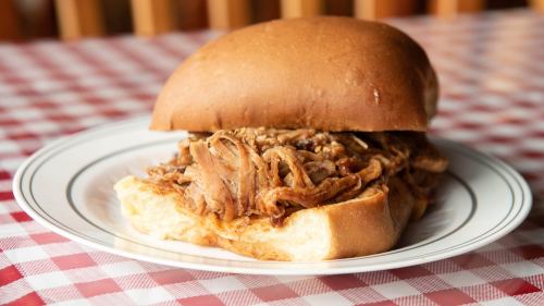 A pulled pork sandwich on a soft bun, served on a plate with a checkered tablecloth in the background.