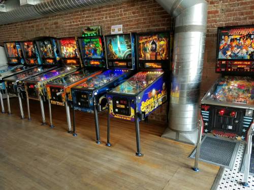 A row of colorful pinball machines lined up against a brick wall in a spacious arcade.