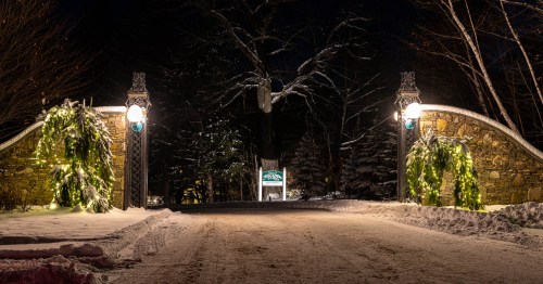A snow-covered entrance with stone pillars adorned with greenery and lights, leading into a dark, wooded path.