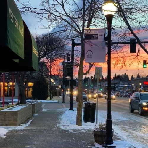 A winter street scene at sunset, featuring a café, traffic lights, and snow-covered sidewalks.