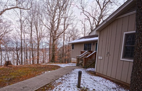 A house with a wooden porch, surrounded by bare trees and a snowy path, overlooking a lake in the background.