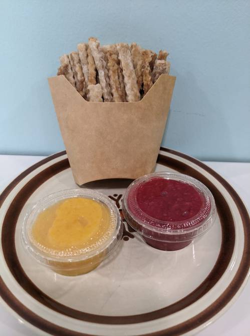A paper container filled with crispy sticks, accompanied by two small cups of yellow and red dipping sauces on a plate.