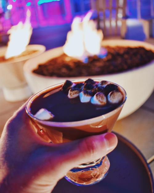 A hand holds a glass of hot chocolate topped with marshmallows, with flames glowing in the background.