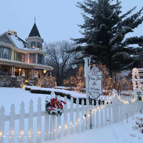 A charming bed and breakfast with festive lights, surrounded by snow and a white picket fence, creating a cozy winter scene.