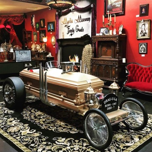 A vintage car designed as a coffin, displayed in a museum with ornate decor and a fireplace in the background.